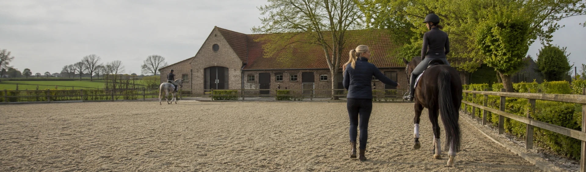 Lesgeefster begeleidt ruiters te paard in de buitenpiste van een Vlaamse manège