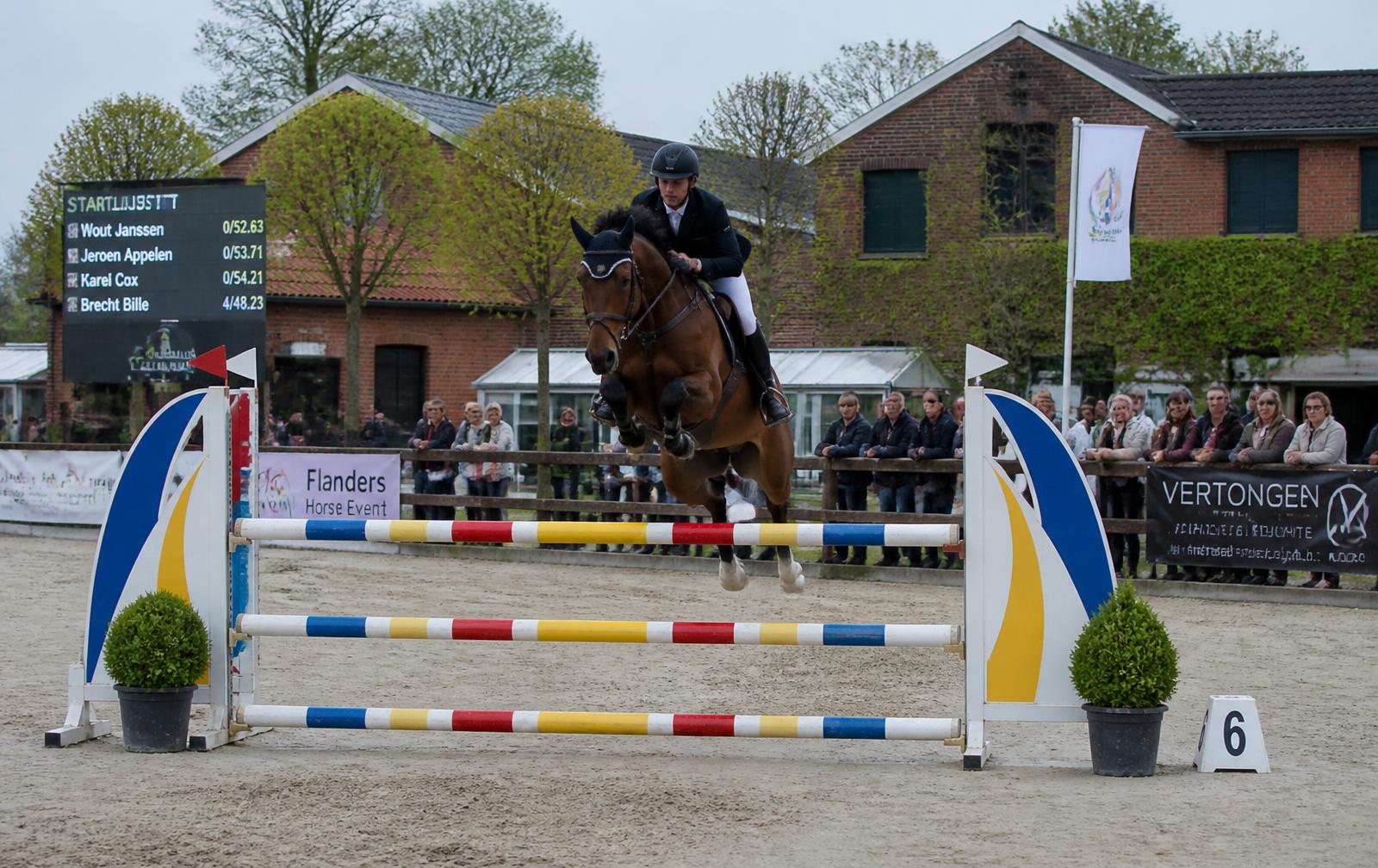 Ruiter springt over een hindernis tijdens een jumpingwedstrijd op een manege met publiek langs de piste