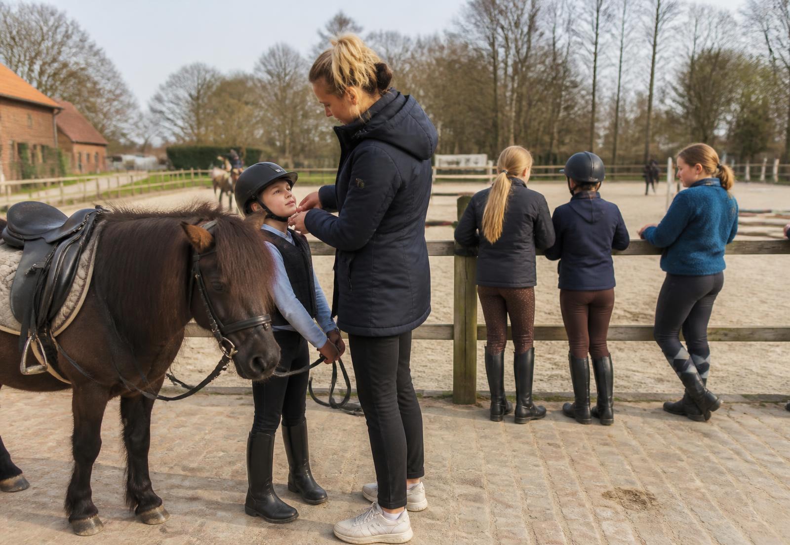 Ouder helpt een kind met de rijhelm naast een pony aan de piste van een manège